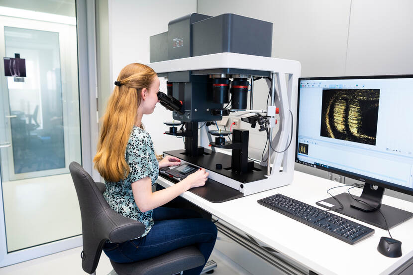 vrouw kijkt via een vergelijkingsmicroscoop naar de hulzen. Naast haar staat een computer met daarop twee gelijkende afbeeldingen.